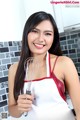 A woman in an apron holding a whisk in a kitchen.