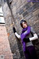 A woman leaning against a brick wall wearing a purple scarf.