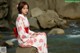 A woman in a red and white kimono sitting on a rock by the water.